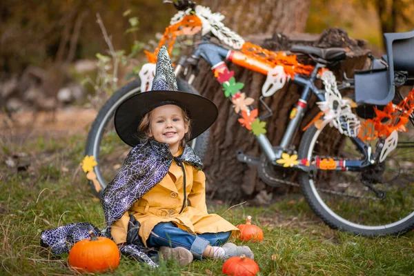 自然の中でハロウィーンの休日に小さな女の子 子供はガーランドで飾られた自転車の近くに座っています — ストック写真