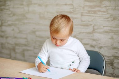 Down sendromlu küçük bir çocuk ahşap bir masada oturuyor ve kağıt üzerinde keçeli kalemle çizim yapıyor. Genetik olarak hasta bir çocuk için eğitim.