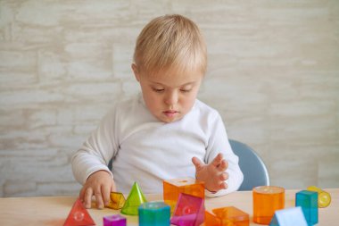 Genetik hastalığı olan küçük sevimli çocuklar masada oturuyorlar. Down sendromlu çocuk 3 boyutlu geometrik figürlerle oynar. Oyuncak içinde plastik.