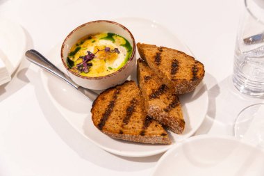 Sıcak, ızgarada pişmiş ekmek dilimleri narin otlarla süslenmiş kremalı yumurta tabağına eşlik ederek sıcak bir kafede cazip bir kahvaltı deneyimi yaratıyor..