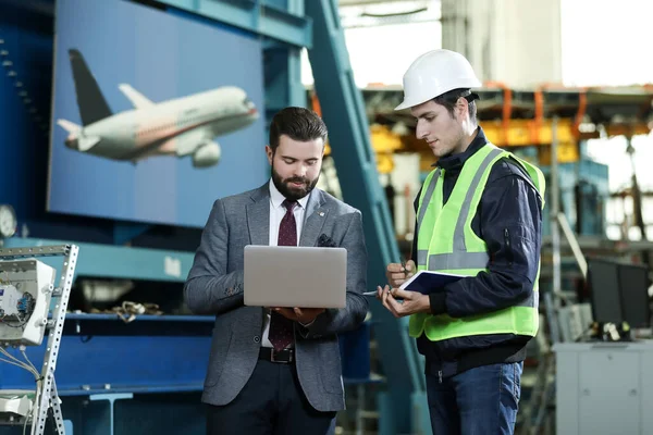 Sağlam bir iş adamının portresi dizüstü bilgisayarı ve fabrika mühendisi uçak fabrikasındaki çalışma sürecini kontrol ediyor..