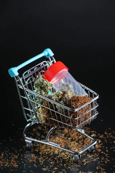 Blue shopping cart full of tobacco and cannabis leafs on a black background. Marijuana legalization. Medical cannabis. Drug addiction.