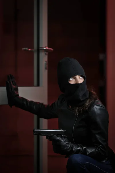 House robbery by woman in a black jacket and black mask holding black gun. Burglar in a mask. Thief in a mask trying to break into other people\'s apartments