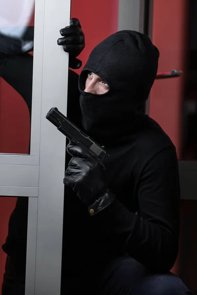 House robbery by man in a black jacket and black mask holding black gun. Burglar in a mask. Thief in a mask trying to break into other people\'s apartments