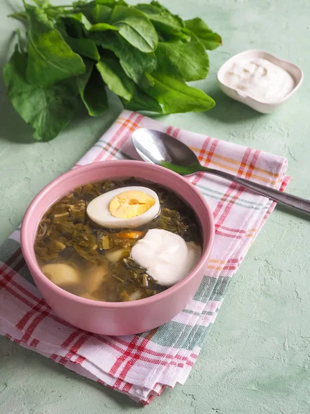 Green soup in a bowl. Sorrel soup with eggs and leaves, onion, bread on a green table.