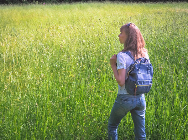 A slender woman with a backpack travels on the tall grass.