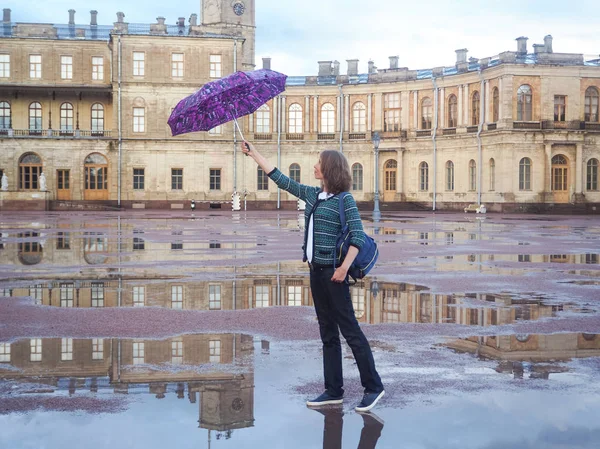 Μια γυναίκα με ένα σακίδιο και ομπρέλα σε ένα πανέμορφο ιστορικό χώρο στο φόντο της ένα αρχαίο παλάτι. Γκάτσινα. — Φωτογραφία Αρχείου