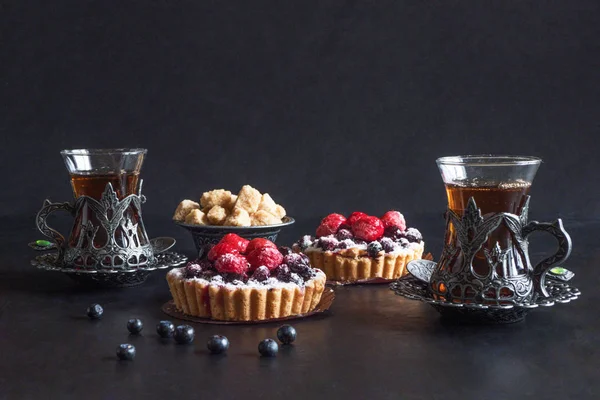 Tea cups, tarts cakes sweets with fresh raspberries and blueberry on black  background — Stock Photo, Image