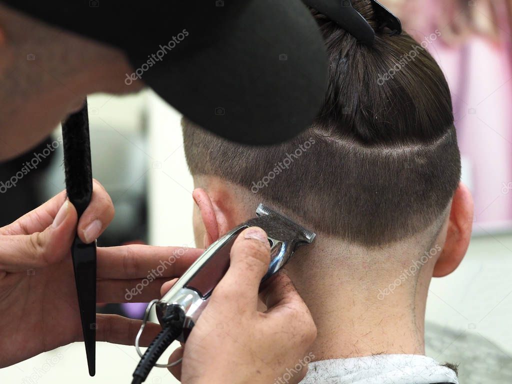 Barber with hair clipper works on haircut of bearded guy barbershop background