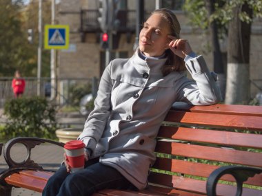 Şehirde açık renk ceketli, orta yaşlı güzel bir kadın yeniden kullanılabilir kahve fincanıyla bankta oturuyor.