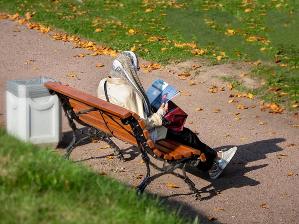 Літня жінка читає, сидячи на лавці з нею назад до міського парку восени — стокове фото