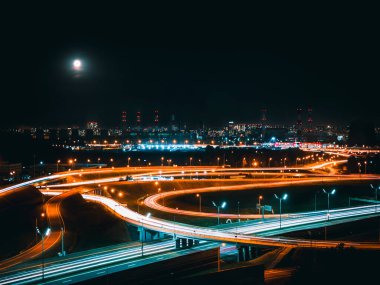 Modern bir metropolün endüstriyel bölgesinde gece yolu kavşağında ay ışığı. Farlardan gelen bulanık ışıklarla uzun deklanşör hızı.