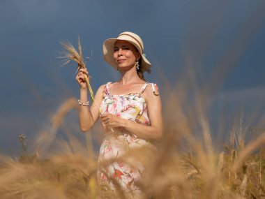 Happy woman in a field with ears. A beautiful and happy woman 49+ in a dress and a hat holds many flowers in her hands against a blue sky backdrop.