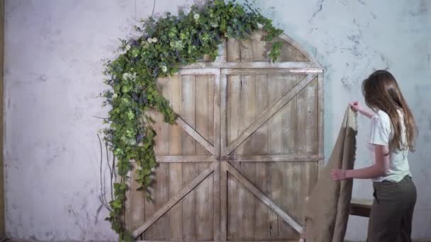 Young girl decorator decorates a staircase with a vintage coverlet on the background of a wooden gate decorated with flowers — Stock Video