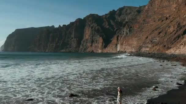 Μια νεαρή γυναίκα περπατάει και απολαμβάνει μια όμορφη θέα στον Ατλαντικό ωκεανό και τα βράχια στη μαύρη παραλία. Λα Γιγάντες, Τενερίφη, Κανάριοι Νήσοι, Ισπανία — Αρχείο Βίντεο