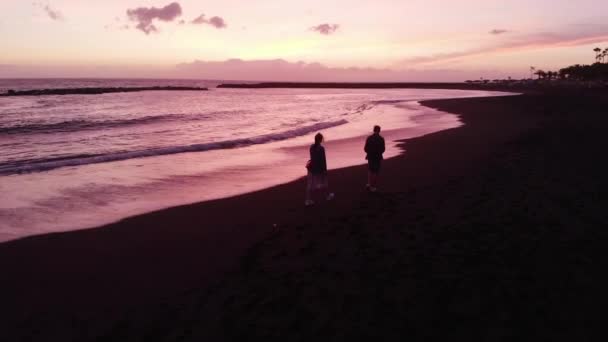 Aérea. Un hombre y una mujer corren a lo largo de una playa volcánica negra en una hermosa puesta de sol rosa. Tenerife, España . — Vídeo de stock