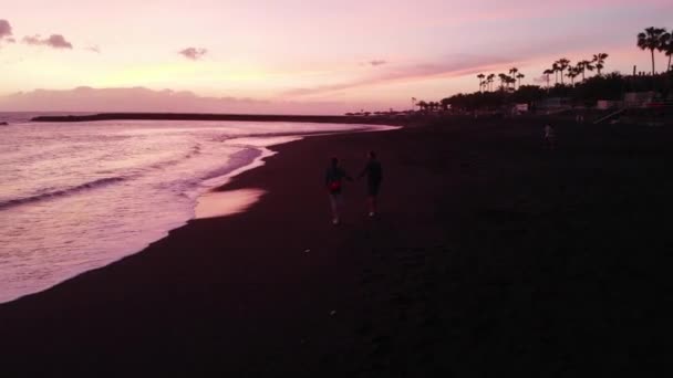 若い家族は日没時に海の黒いビーチに沿って歩き、ドローンからの眺め。スペインのテネリフェ島. — ストック動画