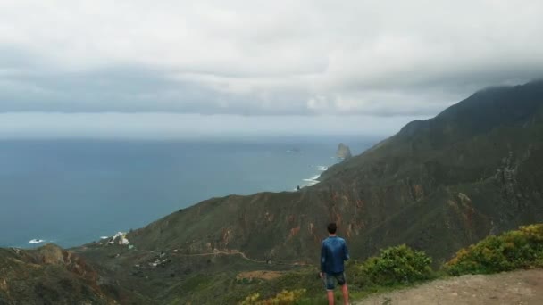 Vista aérea. Un solo hombre se encuentra en el borde de una montaña, mirando una hermosa vista: la larga costa del Océano Atlántico, el valle verde y las montañas del Parque Nacional Anaga, Tenerife — Vídeos de Stock