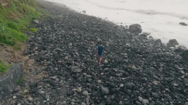 Un homme marche seul le long d'une plage noire faite de grosses pierres. La voie difficile vers l'océan Atlantique. Tenerife, îles Canaries, Espagne. AÉRIAL — Video