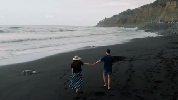 美しい映画のショット。若いカップルが歩いて、黒い火山砂浜に沿って腕で走ります。テネリフェ島、スペインの航空写真 — ストック動画