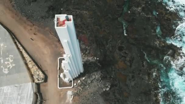AERIAL. Unusual white lighthouse, which stands on the coast of volcanic origin. Lava froze, reaching the ocean. Tenerife, Canary Islands, Spain. Birds eye view — Stock Video