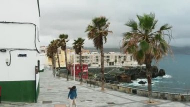 Hava. Sinematik çekim - bir kız ıssız bir İspanyol şehir boyunca yürür, kıyı boyunca, güçlü bir rüzgar sırasında, bulutlu havalarda, bir kuş bakışı görünümünde. Siesta, Gran Canaria, İspanya