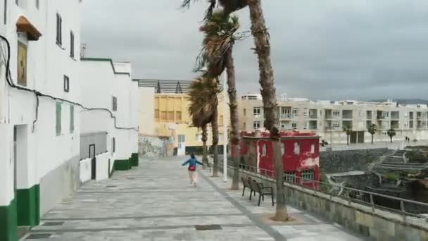 Vista aérea: un avión no tripulado vuela sobre una niña que camina a lo largo de un azulejo antiguo durante un viento huracán, cerca de una pequeña bahía en una isla en el Océano Atlántico . — Vídeo de stock
