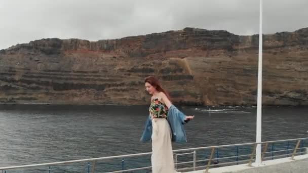 Вид с воздуха - привлекательная девушка прогуливается и сохраняет равновесие на фоне рыбацкой лодки, плывущей по океану в небольшой залив на Канарских островах, Испания — стоковое видео