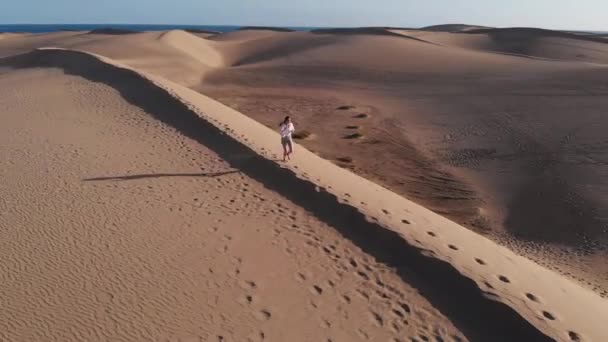 Vista aérea - voo de um drone na frente de uma menina em roupas brancas de verão correndo pelo deserto quente em um dia ensolarado. Maspalomas, Gran Canaria — Vídeo de Stock