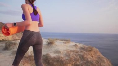 İnce kız spor veya yoga oynamak için bir halı ile bir uçurumun yanına gider. Okyanus ta gün batımı veya şafak.
