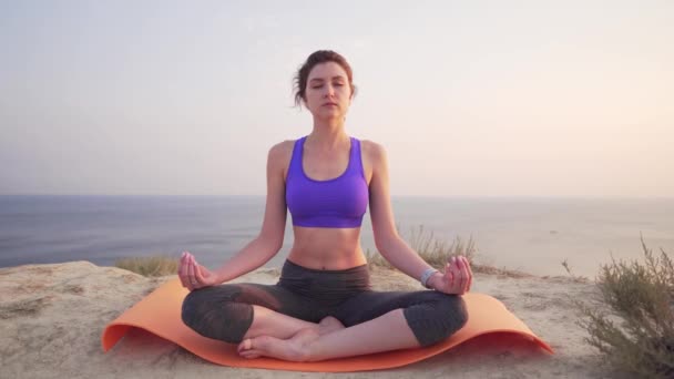 En ung och attraktiv kvinna sitter i en Lotus position, med fokus på andning, vid gryningen vid havet i bergen. — Stockvideo