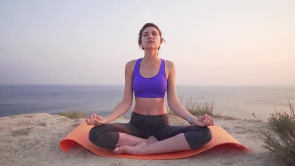 Una bella ragazza si siede in una posizione di loto e alza le mani sopra se stessa, facendo yoga. Atletica donna attraente sullo sfondo dell'oceano . — Video Stock
