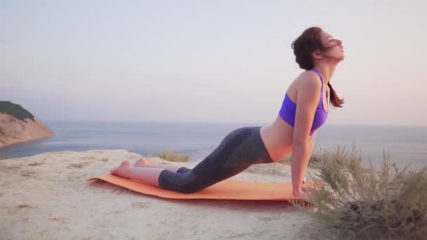 Chica joven y atractiva haciendo estiramiento con vista al mar en el fondo. Mujer relajada y enfocada practicando yoga . — Vídeo de stock