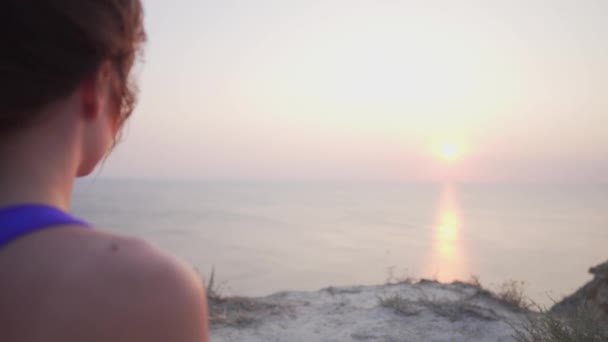 A sports-dressed woman is meditating while watching the sunset on the ocean. View from the back — Stock Video