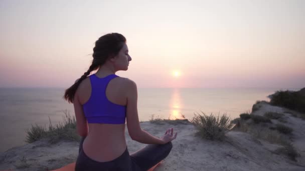 Hermosa y tranquila joven practica yoga con los ojos cerrados - una vista estática desde la espalda, por la mañana junto al océano. Deportes al aire libre — Vídeo de stock