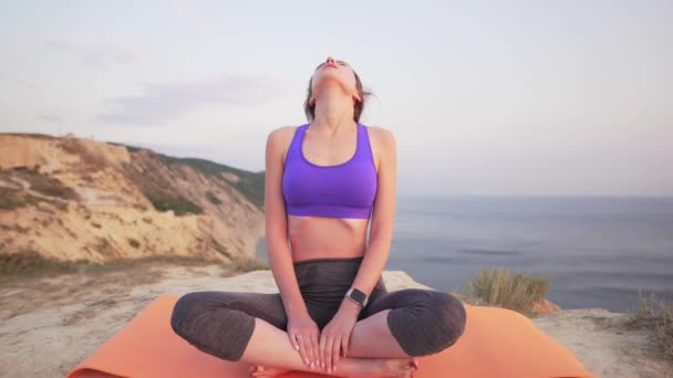 Sports girl practices yoga - focused on breathing, trying to calm herself, mountains and sea in the background, windy day — Stock Video