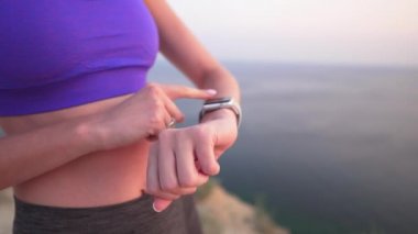 Genç spor kız okyanus kenarında bir uçurumda giyilebilir akıllı saat kullanır. Akşam veya sabah saatlerinde saat ekranına dokunur ve kaydırır. Yakın çekim, saatte bir egzersiz sonuçlarını kontrol