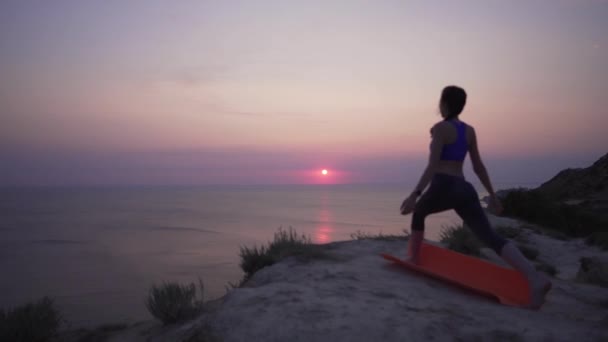 4K Yoga at a cliff above the ocean - a beautifully dressed attractive girl raises her hands to the sky, stands in a yoga pose — Stock Video