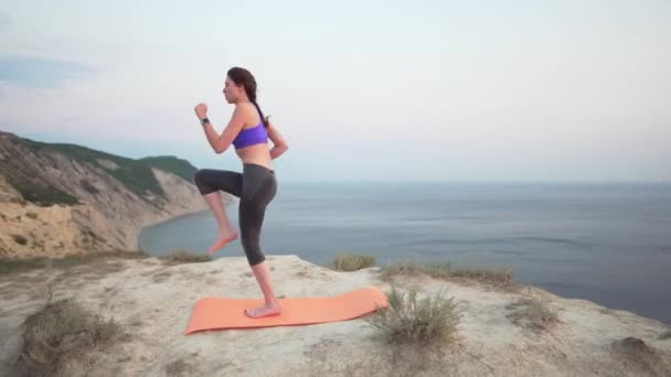 Corredor joven entrenando al amanecer en un acantilado sobre el océano. Preparación para una carrera matutina, atleta en la parte superior y polainas — Vídeo de stock