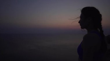 Okyanusun arka planında gün doğumundan önce genç ve beyaz bir kızın silueti, güçlü rüzgar. Yakın çekim meditasyon veya eğitim için hazırlık, sporcu