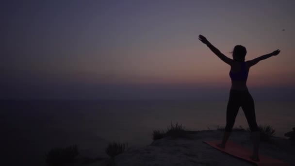 Gün batımında yoga yapan ince genç bir kadının silueti kollarını kaldırıyor. Meditasyon, asana ve doğa ile birlik. Statik çekim — Stok video