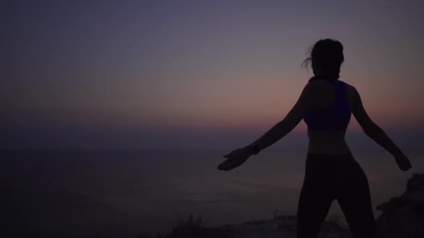 Lugn flicka höjer hennes händer upp som en ballerina, utbildning på naturen. Silhuett av en ung kvinna på bakgrunden av havet och soluppgången — Stockvideo