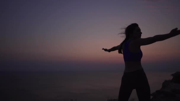 Una ragazza pratica yoga fino all'alba del sole vicino all'oceano in montagna. Asana, braccia ai lati, colpo stabilizzato, concetto di uno stile di vita sano e amore per la natura — Video Stock