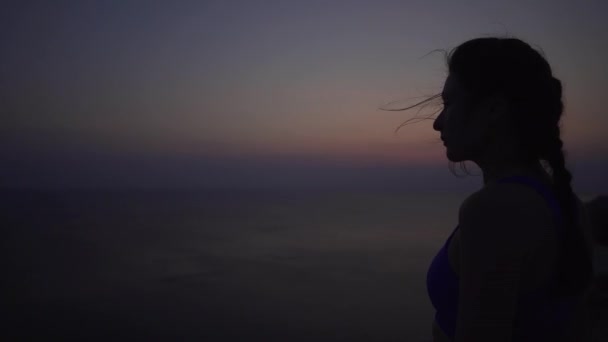 Silhueta de uma jovem bela menina caucasiana antes do nascer do sol no fundo do oceano, vento forte. Close-up de meditação ou preparação para o treinamento, desportista — Vídeo de Stock