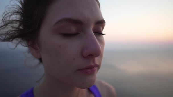 Retrato de cerca de una niña: una atleta con un top púrpura respira antes de meditar, dobla los brazos y practica yoga al atardecer en un acantilado sobre el océano. Largo plazo . — Vídeos de Stock