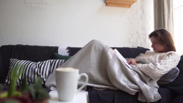 Homeliness - une jeune femme lit un livre sur le canapé, cachée dans une couverture. Concept Hygge — Video