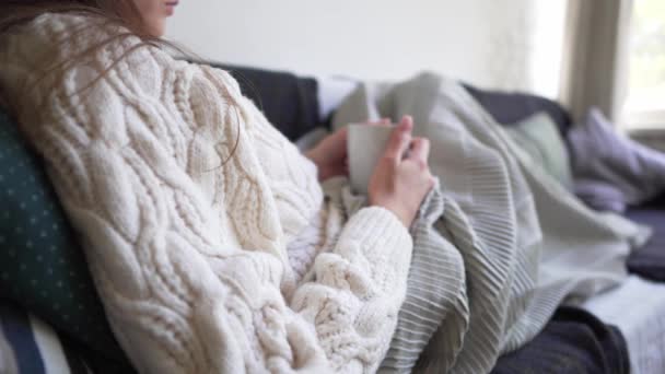 Хигге концепция - женщина в белом свитере держит в руках горячую чашку свежего кофе. Девушка отдыхает на диване, откидывается на подушки и прячется в одеяле — стоковое видео