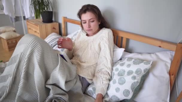 Красивая девушка кладет кофе и открывает книгу, сидя в уютной кровати в деревянном интерьере, прячась в одеяле — стоковое видео