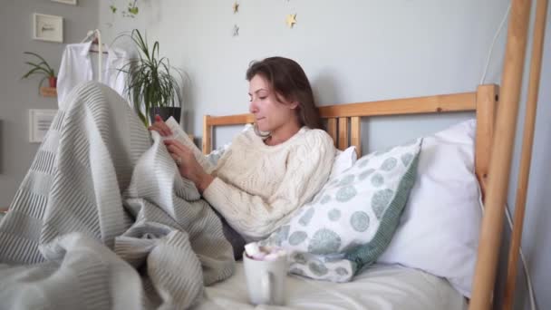 Concept hygge - confort maximum dans son lit dans la chambre à coucher - femme caucasienne lisant un livre et buvant du chocolat chaud un jour d'hiver . — Video