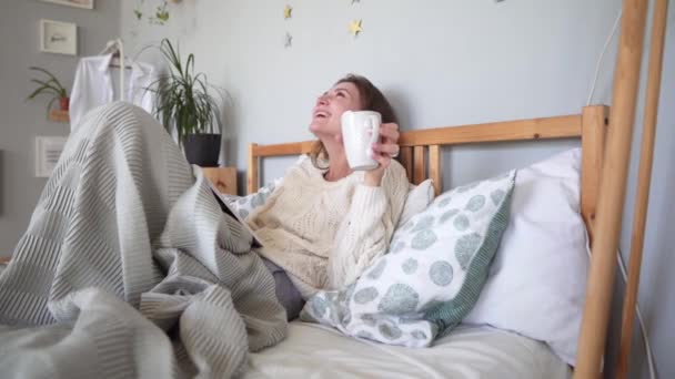 Una hermosa chica atractiva está sentada en la cama, escondida en una manta, leyendo un libro y riendo. Simple felicidad — Vídeos de Stock